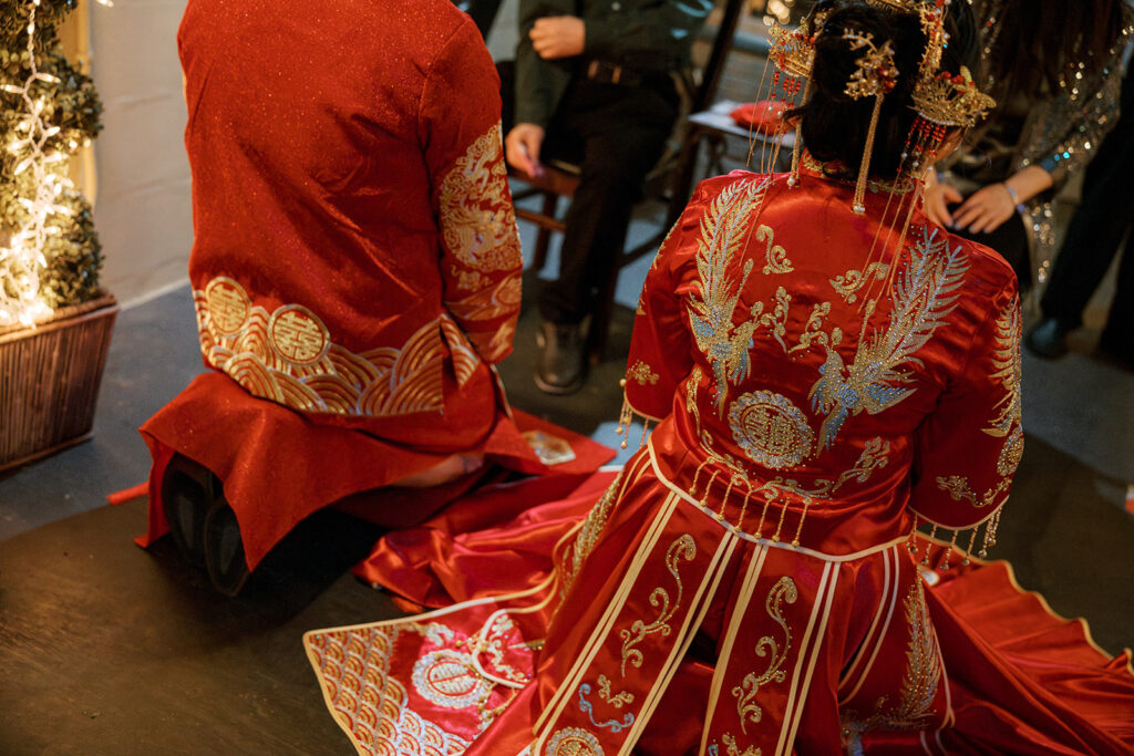 Jessie and Cullen are kneeling together in traditional red and gold outfits during the tea ceremony before their Bryan Museum garden wedding.