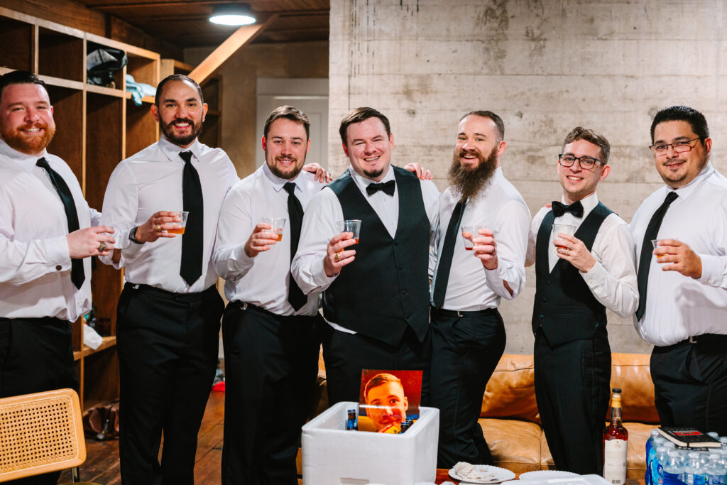 Groom and groomsmen raising glasses together while preparing for a wedding at The Artisan.