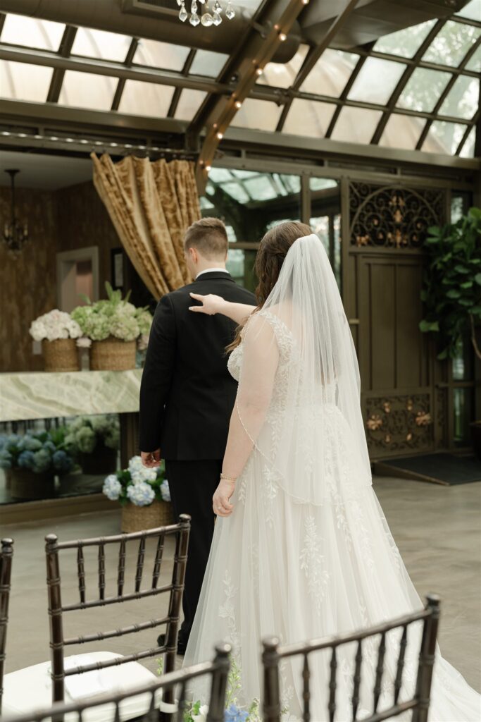 Bride taps groom's back during the first look in the elegant glass conservatory at The Bryan Museum.