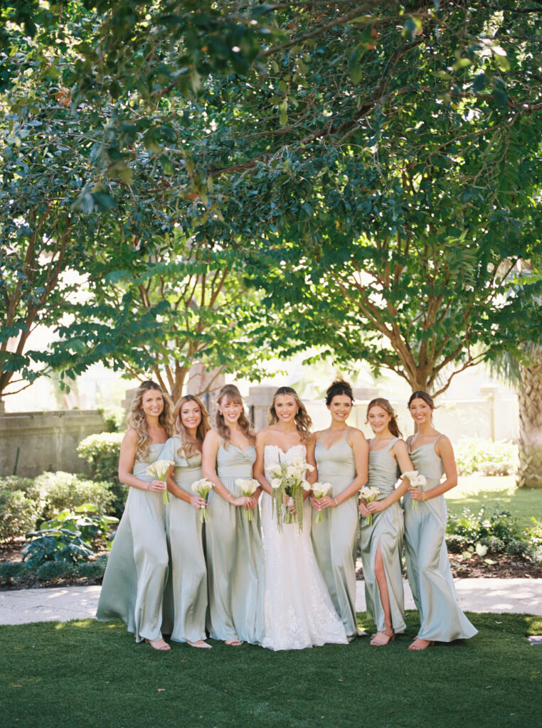Bride and bridemaids smiling outdoors beneath trees during romantic fall wedding portraits at The Bryan Museum.