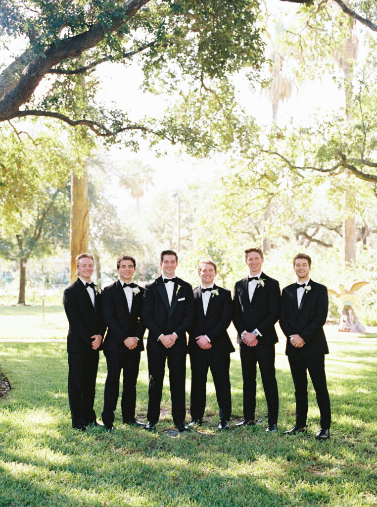 Groom and groomsmen smiling outdoors beneath trees during romantic fall wedding portraits at The Bryan Museum.