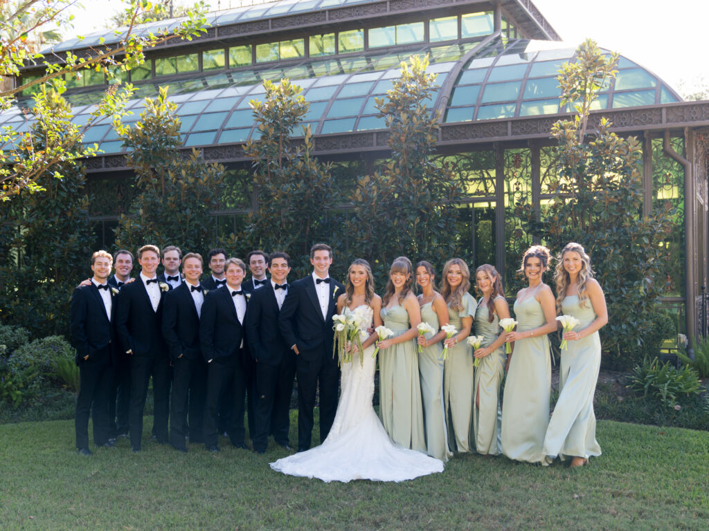Bride, groom, and whole wedding party smiling outdoors by the Bryan Museum Conservatory during romantic fall wedding portraits.